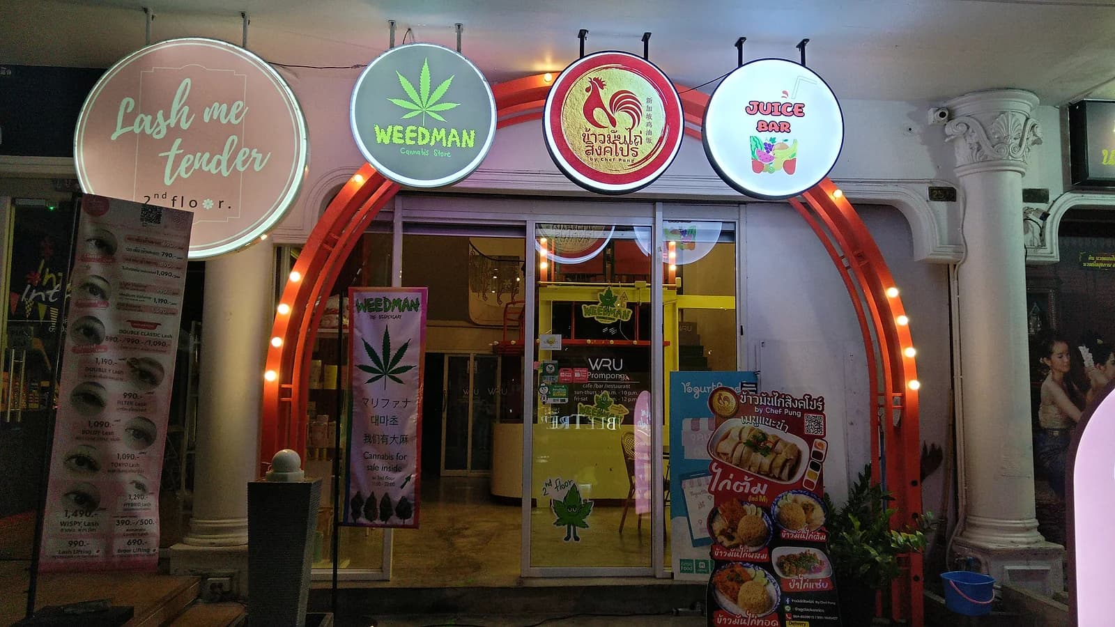 Weedman, a Bangkok cannabis store, sharing a storefront arch with a juice bar and an eyelash studio. The license requirements that followed the 2025 crackdown pushed many smaller, informal sellers out of business.